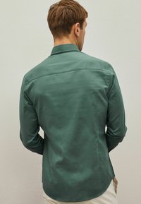 Zöld gombos ing sima anyagból, enyhe fényességgel, klasszikus gallérral és hosszú ujjakkal, ívelt szegéllyel.