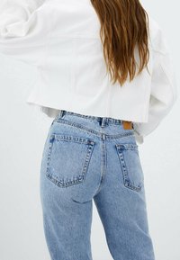 Bílá zkrácená bunda přes světle modré džíny s vysokým pasem. Džíny mají vybledlou texturu se dvěma zadními kapsami a šitými detaily.