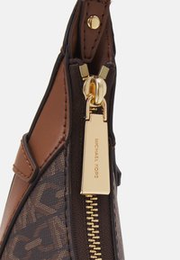 Καφέ δερμάτινη τσάντα χειρός με χρυσό φερμουάρ και ετικέτα. Το σώμα της έχει μονογράμματα με σκούρες καφέ λεπτομέρειες και λεία υφή.