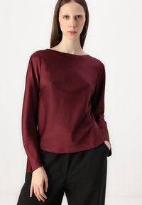 Burgundy long-sleeve top with a boat neckline. Smooth satin material, loose fit, and side slits. Paired with black, loose-fitting pants.