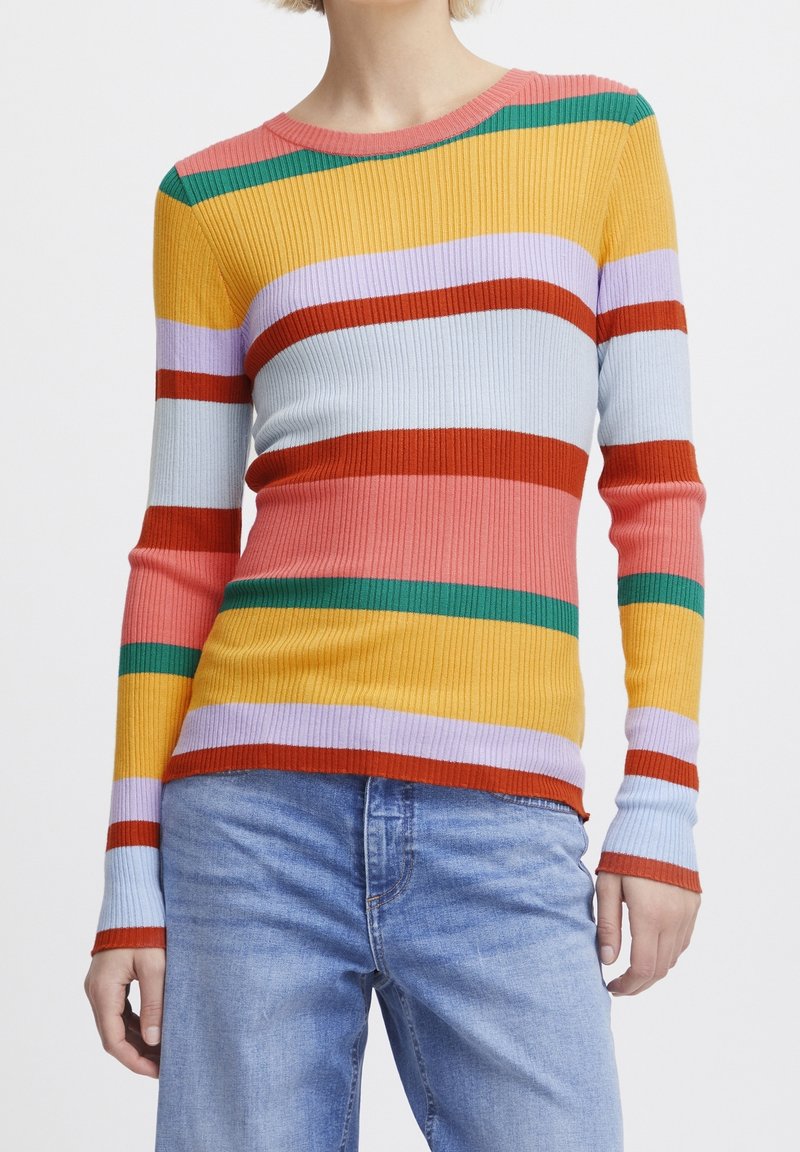 Femme portant un pull côtelé à manches longues avec des rayures horizontales rose, vert, jaune, rouge et bleu clair, associé à un jean bleu clair.