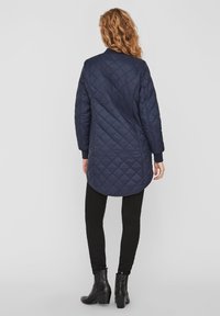 Kviltad lång kappa i marinblått med rundad nederkant, ribbade ärmslut och en slät yta. Bakdelen har diagonala sömmar för extra detalj.