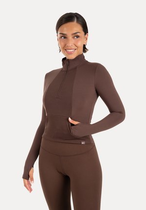 Kvinde iført en brun langærmet, tætsiddende atletisk top med halvt lynlås og matchende leggings, smilende og poserende med den ene hånd i en lomme.