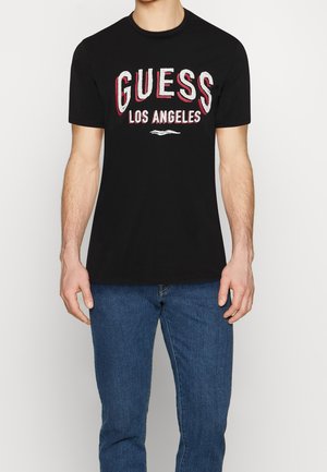 Mężczyzna ubrany w czarną koszulkę z krótkim rękawem z napisem "GUESS LOS ANGELES" w białym i czerwonym kolorze, w połączeniu z niebieskimi dżinsami na jednolitym tle.