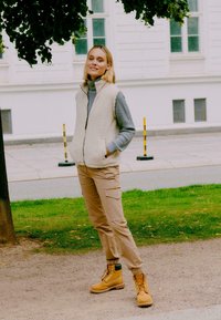 Gilet in pile beige sopra un maglione grigio, pantaloni cargo color beige e stivali gialli. Il gilet ha una chiusura con zip e tasche laterali, ambientato all'aperto.