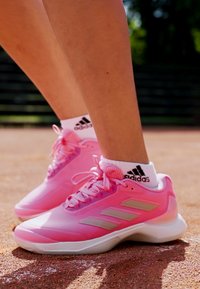 Pink athletic shoes with a synthetic upper, featuring three silver stripes and a thick white sole, paired with white socks bearing a logo.