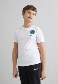 Chlapec v bielom tričku Nike a čiernych teplákoch Nike stojaci pred obyčajnou bielou textúrovanou stenou, pozerá sa mierne doprava.