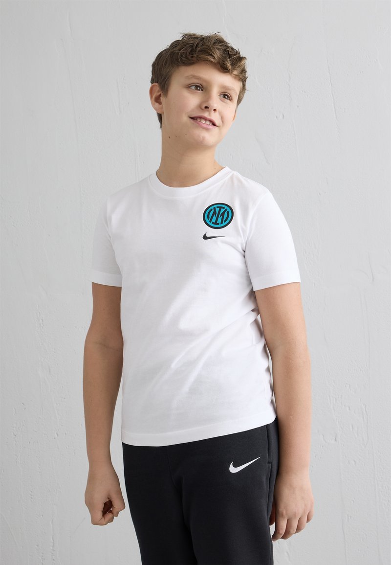 Chlapec v bielom tričku Nike a čiernych teplákoch Nike stojaci pred obyčajnou bielou textúrovanou stenou, pozerá sa mierne doprava.