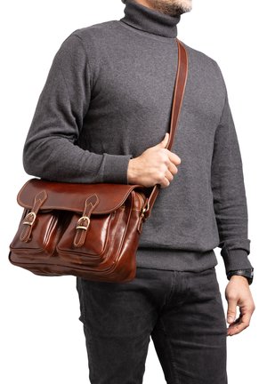Borsa a tracolla in cuoio marrone con due tasche frontali, ferramenta in ottone e una lunga tracolla regolabile, indossata a tracolla sopra un maglione grigio.