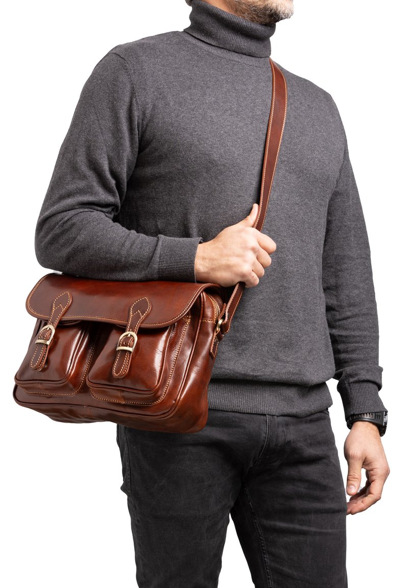 Borsa a tracolla in cuoio marrone con due tasche frontali, ferramenta in ottone e una lunga tracolla regolabile, indossata a tracolla sopra un maglione grigio.