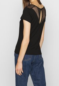 Černé tričko krátkého rukávu s strukturovaným síťovaným vsadkem na zadní straně, s detailem ve tvaru klíčové dírky a volným střihem. Nosené s modrými džíny.