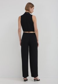 Femme aux cheveux courts portant un crop top noir sans manches, un pantalon noir taille haute, des boucles d'oreilles en argent et des sandales à talons noirs, de dos.