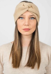 Il cerchietto intrecciato beige è realizzato con un design frontale a torsione, ha una consistenza morbida ed è dotato di una vestibilità elastica. Indossato con un maglione beige chiaro.