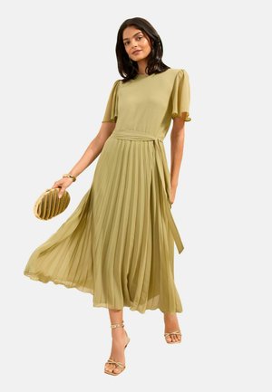 Frau trägt ein hellolivgrünes, plissiertes Midikleid mit kurzen Flatterärmeln, hält eine goldene Clutch und trägt goldene Riemchenabsätze.