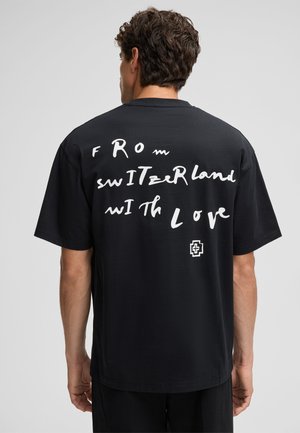 Black cotton t-shirt featuring white handwritten text "FROM SWITZErLand WITH Love" on the back and a small logo in white at the lower left.