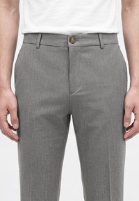 Homme portant un pantalon gris ajusté avec fermeture par bouton et zip, accompagné d'une chemise blanche rentrée, mains détendues le long du corps.