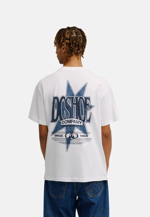 Osoba z krótkimi twistami ma na sobie białą koszulkę z dużą niebieską gwiazdą i napisem "DC SHOE COMPANY" z tyłu, zestawioną z niebieskimi dżinsami.