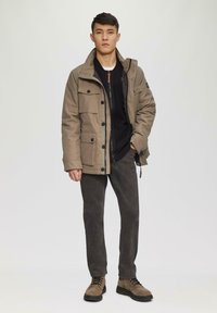 Tan gewatteerde jas met meerdere voorzakken, zwarte binnenvoering, gecombineerd met donkergrijze broek en bruine veterschoenen. Eenvoudig, functioneel ontwerp.