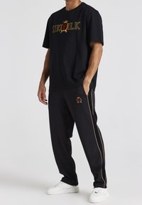 Czarny bawełniany T-shirt z wyszywanym na złoto logo "SIKSILK", w połączeniu z czarnymi joggerami z boczną lamówką i czerwoną koroną. Białe sneakersy.