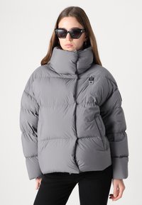 Fyldig grå jakke med høj krave, quiltet design og lomme detaljer. Båret med sorte solbriller og mørke bukser, sat mod en ensfarvet baggrund.