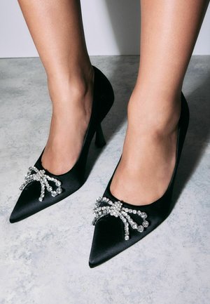Escarpins noirs en satin avec un design à bout pointu, ornés d'un nœud embellissant en cristaux sur le coup de pied et de talons aiguilles fins.