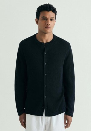 Joven con cabello corto y rizado lleva un cárdigan negro abotonado y pantalones blancos, de pie frente a un fondo claro y sencillo.