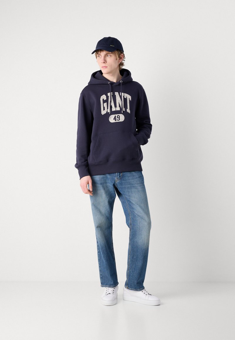 Felpa navy con "GIANT 49" stampato in bianco, abbinata a jeans leggeri blu e sneakers bianche; un cappellino navy che completa l'outfit.