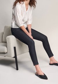 Femme portant un chemisier blanc, un jean skinny foncé et des chaussures noires pointues sans lacets, assise sur un fauteuil gris clair avec des pieds noirs.