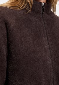 Dark brown zip-up jacket made from soft, textured fabric with a high collar and minimalistic design. Features contrasting zipper hardware.