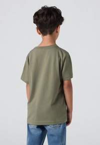 Alyvuogių žalios spalvos medvilnės marškinėliai trumpomis rankovėmis, su apvaliu iškirpimu ir laisvu siluetu, derinami su šviesiai mėlynomis džinsinėmis kelnėmis. Paprastas dizainas, be raštų.