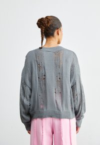 Grå stickad tröja med stora öppna stickade sektioner och en avslappnad passform, med långa ärmar och ribbat nederkant, parat med rosa byxor.