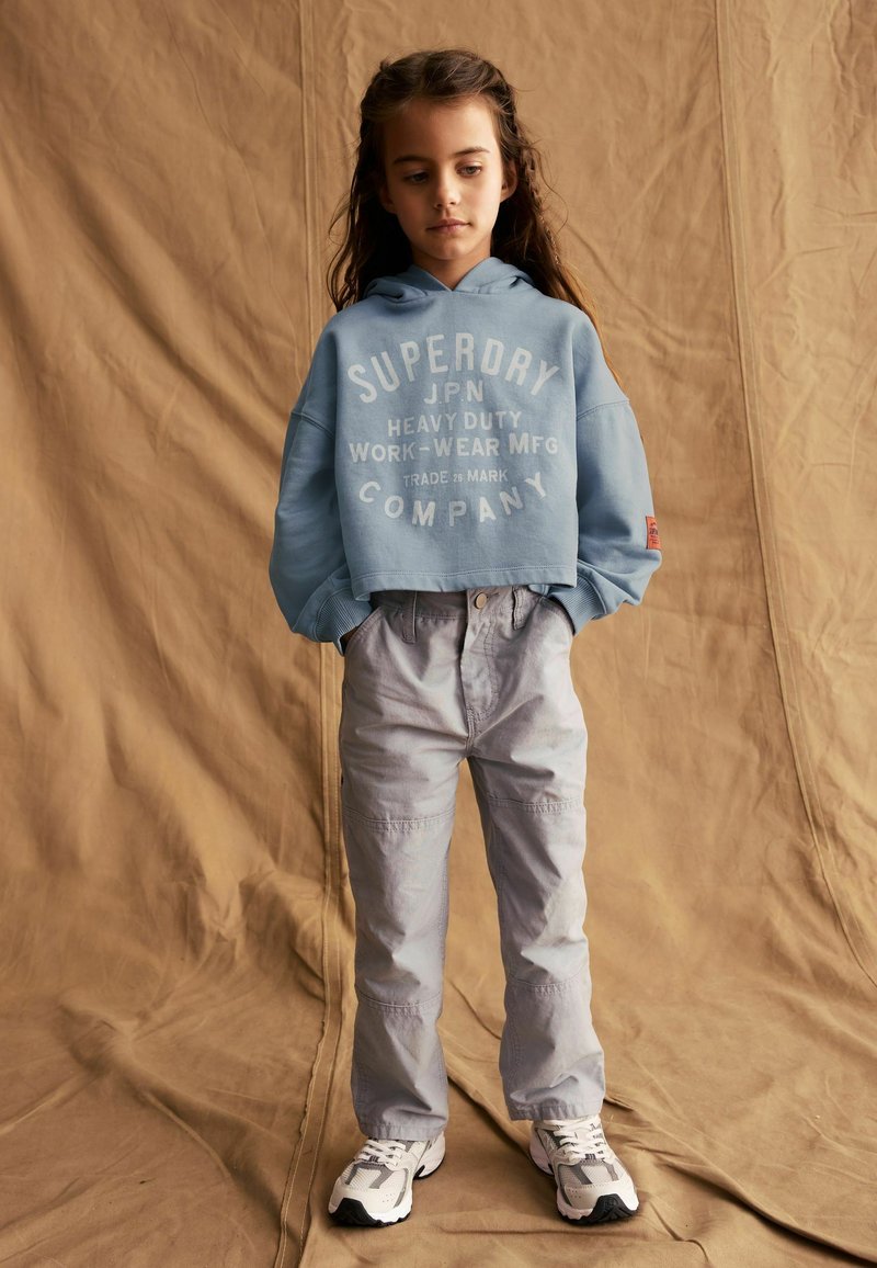 Felpa cropped azzurra con marca in bianco; pantaloni cargo grigi a fit largo; scarpe da ginnastica bianche e grigie. Sfondo neutro.