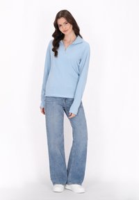 Suéter azul claro de manga larga con cuello de cremallera, combinado con unos jeans de mezclilla de lavado claro de corte holgado y zapatillas blancas.