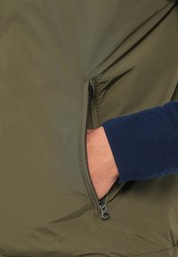 Μπουφάν σε ελιά πράσινο με υφή στην εξωτερική επιφάνεια, που διαθέτει τσέπη με φερμουάρ και μανσέτα σε παλτό μπλε χρώμα που εκτείνεται από το μανίκι.