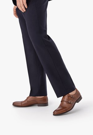 Hombre con pantalones azul marino y zapatos de cuero marrones con doble hebilla, de pie con un pie ligeramente levantado.