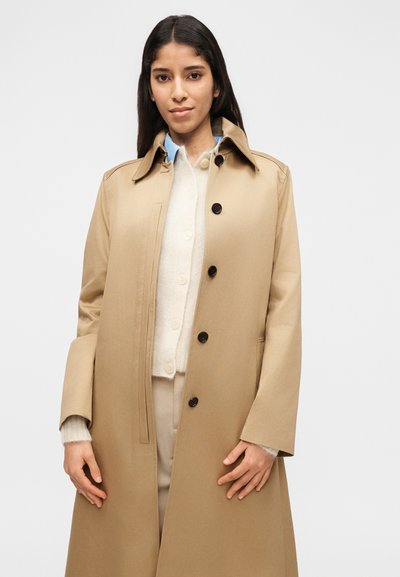 Γυναίκα με μπεζ παλτό τρεντζ με κουμπιά πάνω από λευκή ζακέτα και γαλάζιο πουκάμισο με γιακά, που στέκεται μπροστά σε απλό λευκό φόντο.