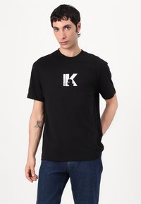 Juodas medvilninis marškinėlis su apvaliu kaklu ir trumpomis rankovėmis, su dideliu baltu grafiniu logotipu krūtinėje. Kasdienis siluetas.