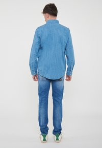 Camicia in denim azzurro chiaro con colletto a punta, maniche lunghe e orlo arrotondato, abbinata a jeans azzurro chiaro e sneaker bianche con dettagli verdi.