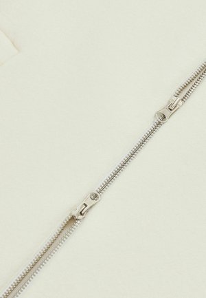 Fermuar din metal argintiu cu dinți netezi, așezat pe un fundal de țesătură de culoare crem. Două tab-uri pentru operare ușoară.