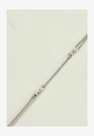 Reißverschluss aus silbernem Metall mit glatten Zähnen, vor einem cremefarbenen Stoffhintergrund. Zwei Ziehknöpfe für eine einfache Handhabung.