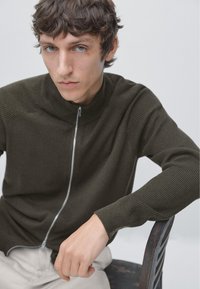 Dark green, zip-up sweater with ribbed texture and high neck. Fitted sleeves, worn by a seated person, hands resting on a chair.