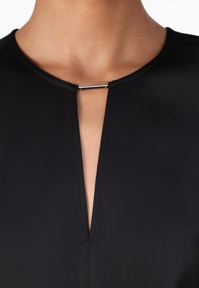 Czarna bluzka z dekoltem w serek, z srebrnym dekoracyjnym zapięciem przy kołnierzyku, prezentująca gładką strukturę materiału i czyste linie.