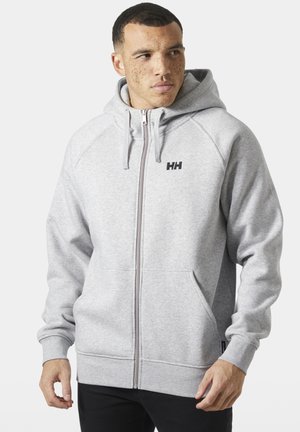 Hombre con cabello corto y pecas lleva una sudadera con capucha gris claro de Helly Hansen con cremallera y bolsillos delanteros, de pie frente a un fondo liso.