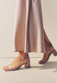 Sandali lilla con tacco a blocco, punta quadrata e cinturino alla caviglia, abbinati a una gonna fluida di raso rosa chiaro. Texture liscia, design semplice.