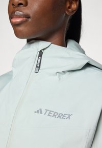 Lys mintgrøn vandtæt jakke med høj krave, lynlåslukning og et grå "TERREX" logo på brystet. Glat tekstur, minimalistisk design.