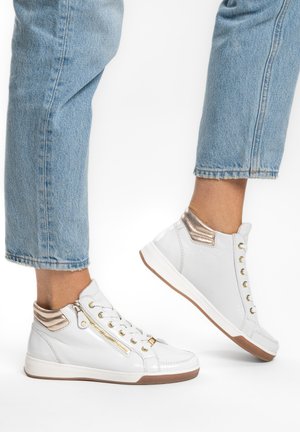 Person wearing white high-top sneakers with gold accents and light blue cuffed jeans, one foot slightly lifted against a white background.