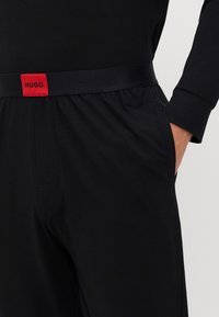 Pantalones deportivos negros con una textura suave, que cuentan con una ancha cinturilla elástica con una prominente etiqueta roja de "HUGO" y bolsillos laterales.