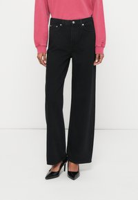 Széles szárú fekete farmernadrág magas derékkal, ötlovas zsebes kialakítással, kontrasztos varrással és sima textúrával, fekete magassarkúval párosítva.