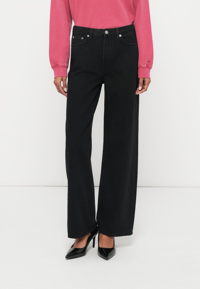 Széles szárú fekete farmernadrág magas derékkal, ötlovas zsebes kialakítással, kontrasztos varrással és sima textúrával, fekete magassarkúval párosítva.