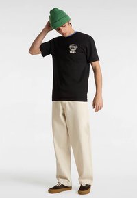 Czarny bawełniany t-shirt z nadrukiem, beżowe szerokie spodnie, brązowe buty sznurowane oraz zielona dziergana czapka. Codzienny strój.